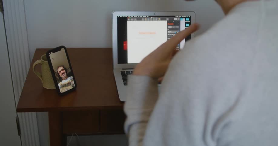 Man sitting in front of laptop signs to man shown in video call on smartphone propped against coffee cup