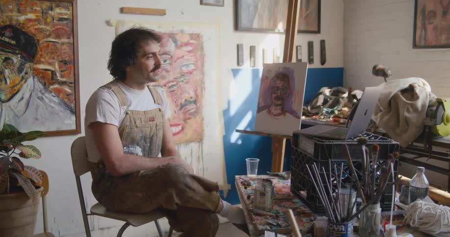 A man is sitting in a chair surrounded by his artwork that he painted