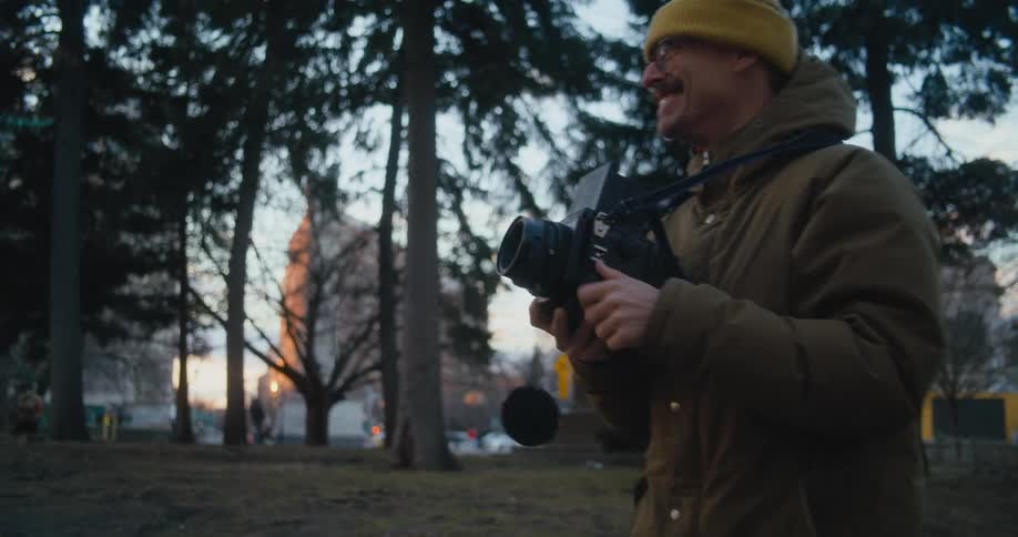 Man wearing a toque and a winter coat takes a photo with a camera in a park