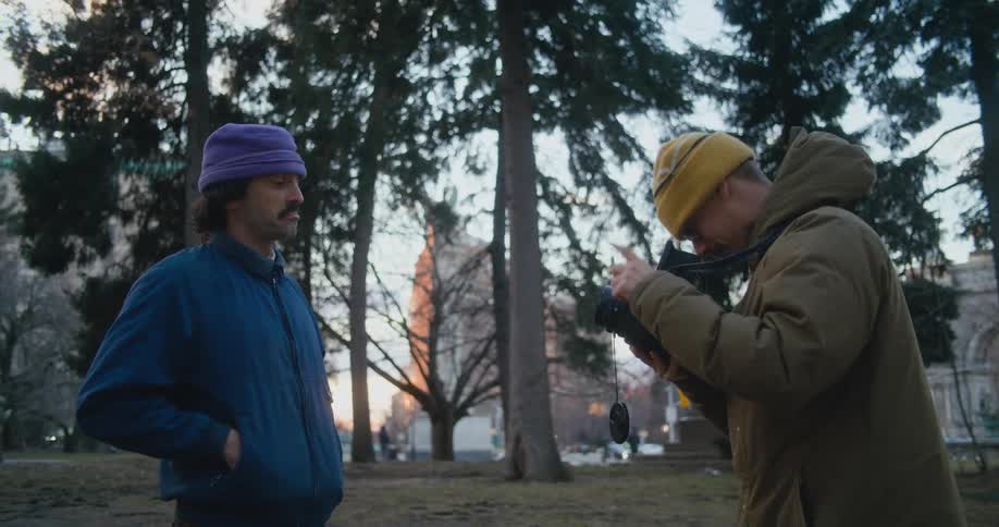 Two men are standing near trees in a park and one man is holding a camera speaking to the other man using sign language