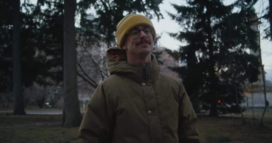 Man with yellow wooly hat glasses mustache and green coat talks to person using sign language in public park
