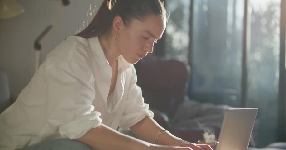 A woman sitting thinking and looking down at her laptop begins to type while steam rises from the mug beside her
