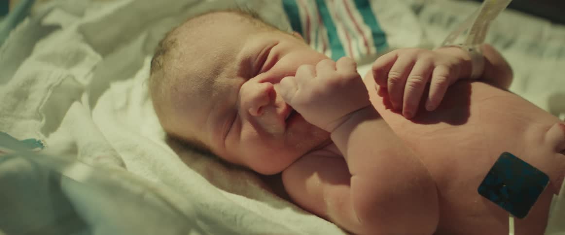 Somebody wearing blue rubber gloves is standing over a newborn baby hooked up to tubes in a hospital bed