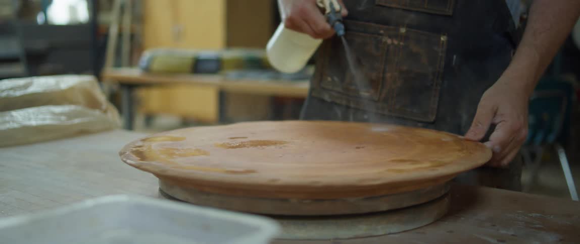 Person standing in pottery studio sprays water onto turntable then wipes it with wet sponge
