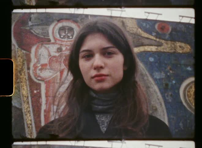 A vintage film with a teenage girl standing outside in front of a mural