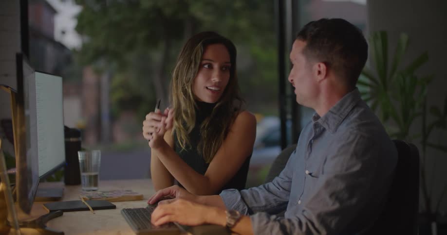 A woman and man talk while sitting together at a computer desk in the evening
