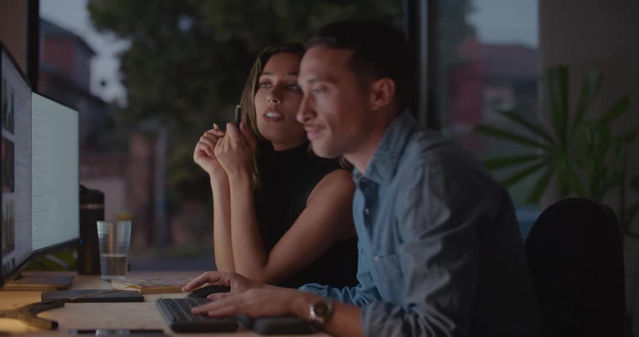 A woman points at a computer monitor while speaking to a man operating the computer while sitting next to her
