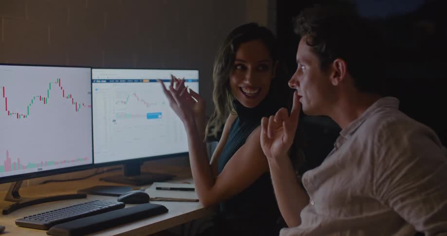 A man and woman sitting together at a desk chat while looking at charts displayed on computer monitors