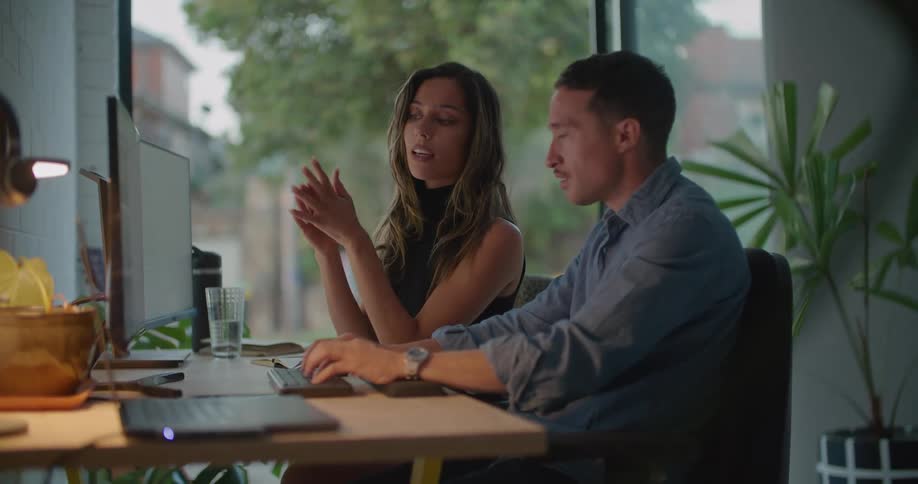 Coworkers sit at desk discussing contents of computer screen as car passes on nearby street