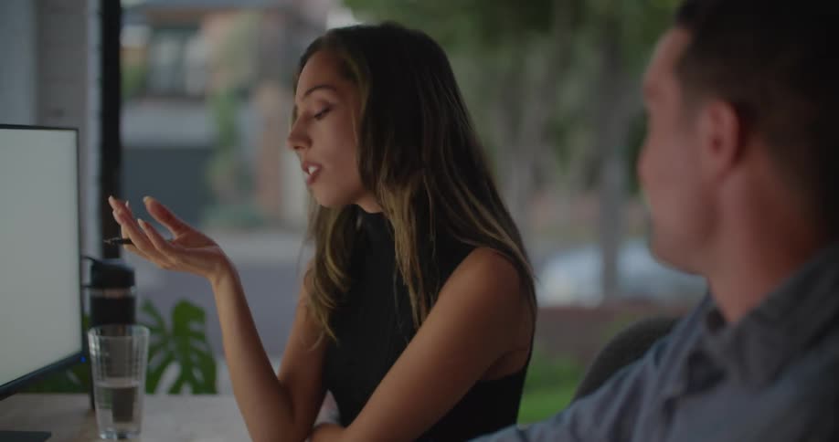 A man and woman sit at a desk and talk in front of computers as they brainstorm in an office