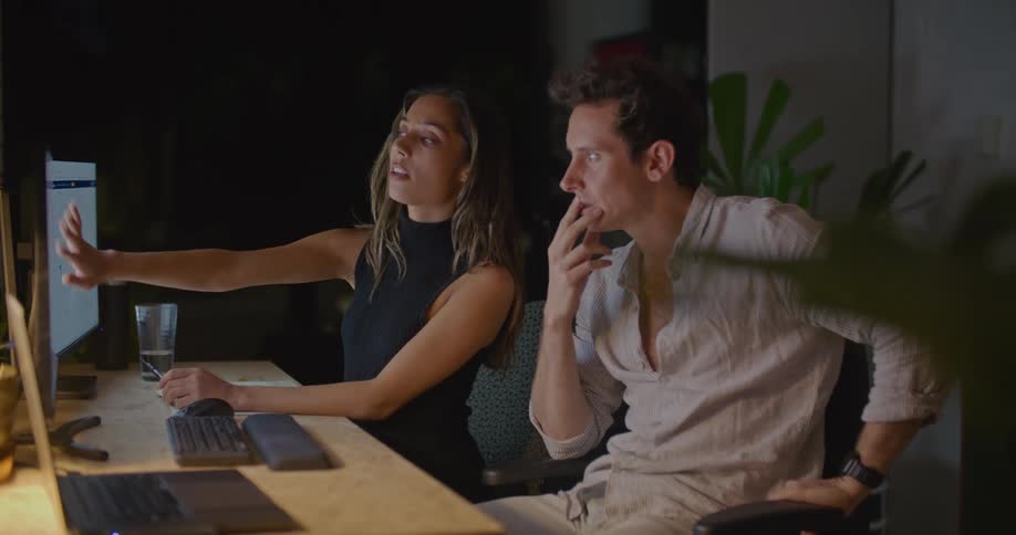 A man and woman sit at a desk in an office and brainstorm as they talk and point at computer monitors
