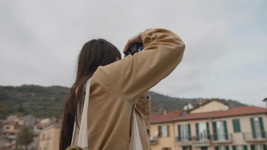 A photograph raises her camera to take photographs of her surroundings while standing outdoors in a city on a cloudy day