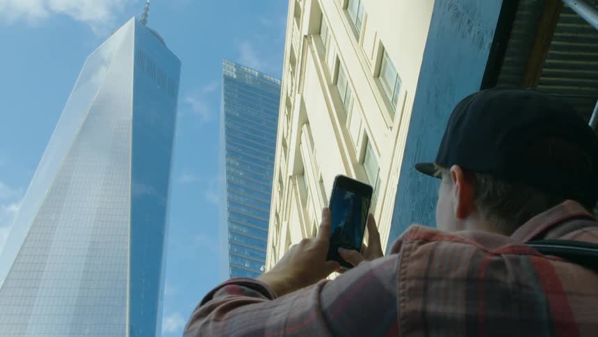 Man wearing a cap and a plaid shirt uses a cellphone to take a photo of a one world trade center on a sunny day