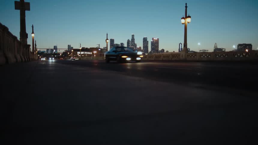 Headlights on cars shine as they travel on a road near downtown and tall buildings and skyscrapers are in the distance