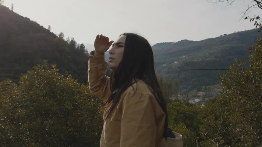 A woman with long brown hair shields her eye from the sun while looking around at the nearby mountains