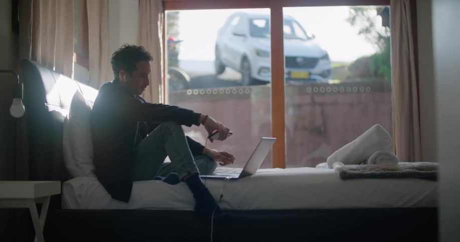 A man sits on his bed indoors with his laptop while conversing on his phone with an earpiece