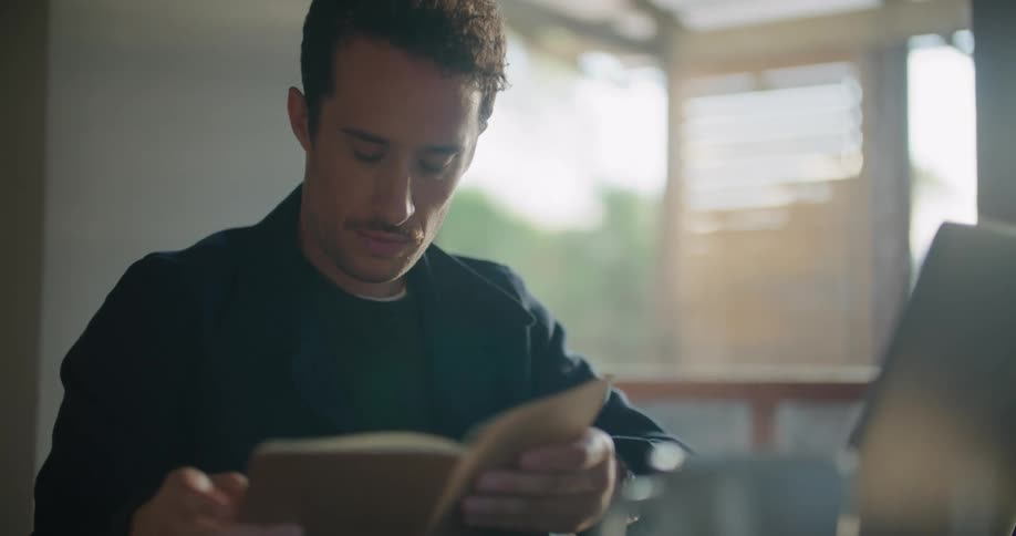 Man with dark jacket checks notebook and works on laptop