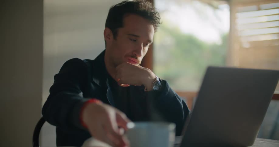 A man in a room sitting on a chair at a table and using a laptop