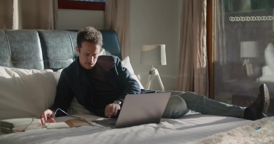 A man stands contemplatively outdoors for a moment before entering a bedroom through a glass door and writing in a notebook while glancing at the screen of a laptop