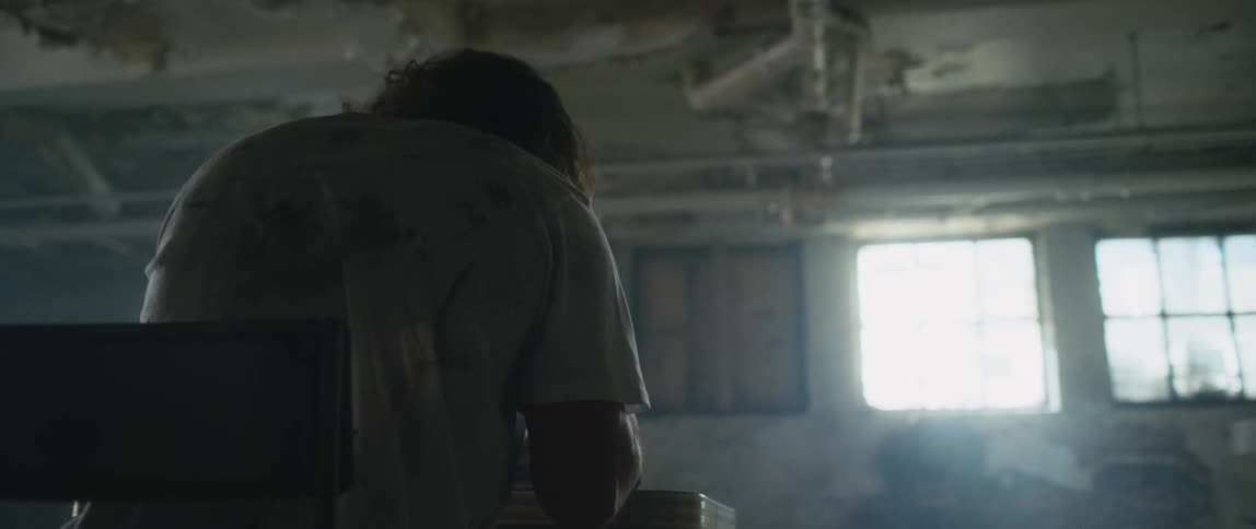 A man wearing a dirty t shirt sitting at a desk flipping through a book in an abandoned school