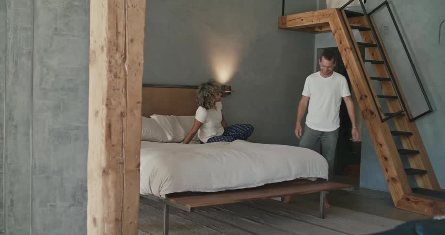A woman sits on a bed texting and a man comes in the room and they talk and the woman gets up and walks out of the room as the man walks further into the room