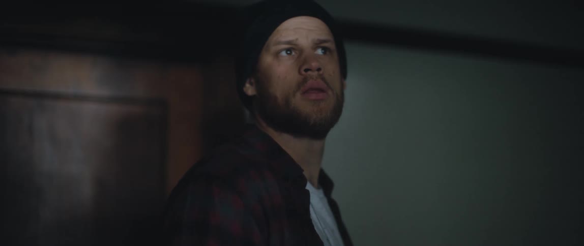 A man wearing a knit cap is walking down a hallway and looking around in a suspicious manner