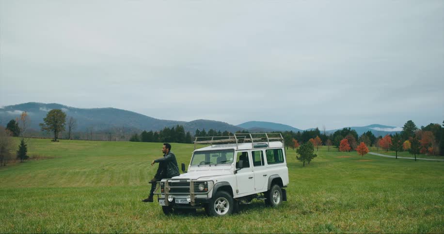 Man sits on hood of his car parked in field in front of mountains