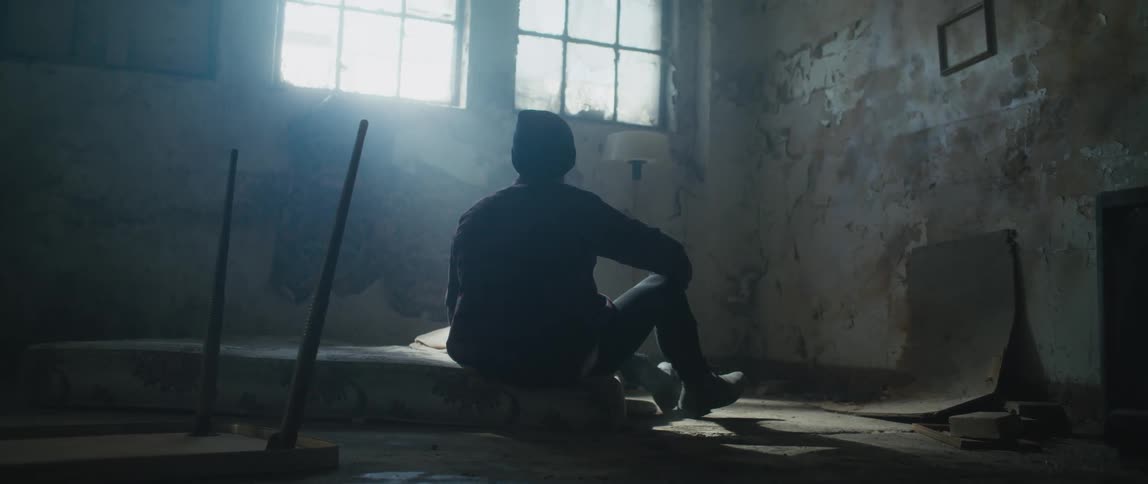 Man sits on mattress on the floor in dimly lit room with peeling paint