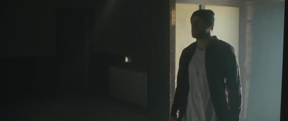 A man wearing a flannel shirt and a knit cap opens a door to a hallway and walks into a dark room