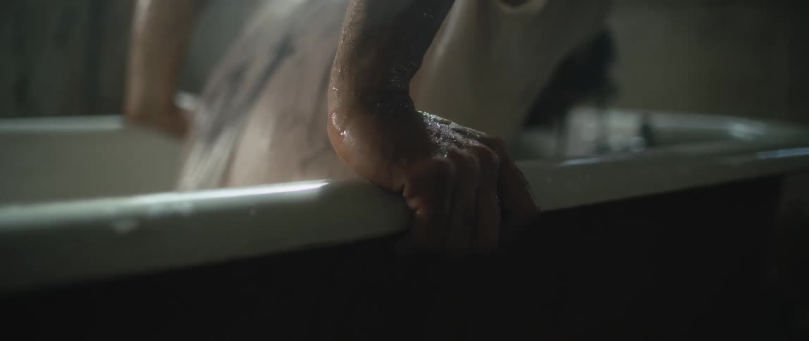 Water splashes as a person wearing a white shirt emerges from a bathtub holding the sides of the tub with their hands