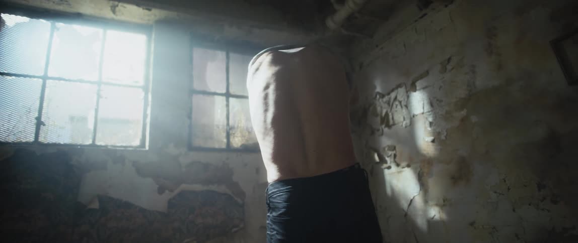 A man is standing in a dilapidated room and he takes off his dirty shirt and angrily throws it on the floor