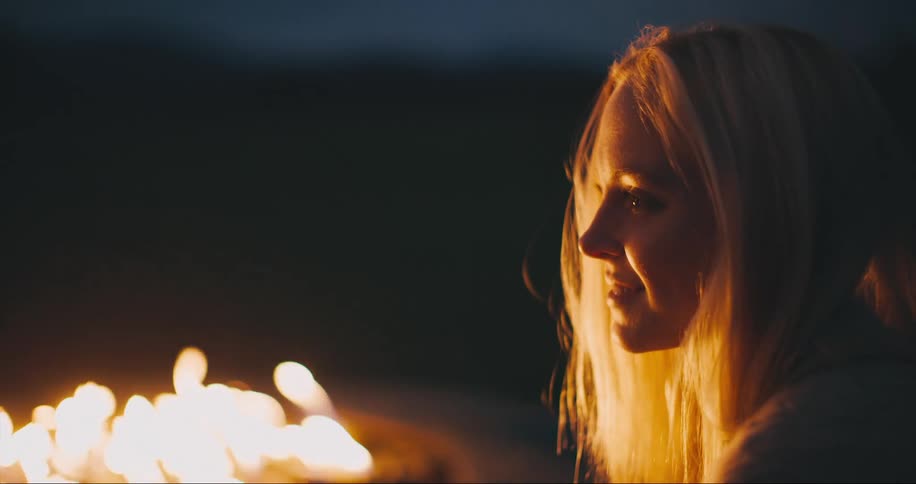 Blonde woman sits by bonfire smiling at night