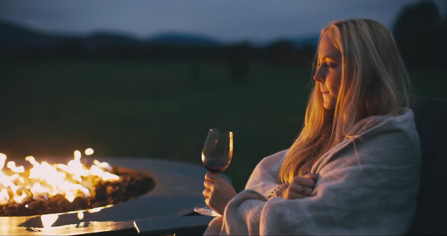 Woman with blanket drinks  glass of wine next to bonfire at dusk