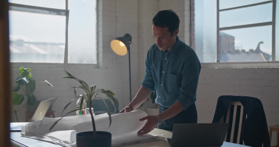 A man in a room standing in front of a desk with a laptop with a wall and windows in the background
