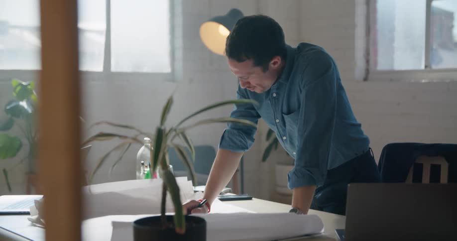 A man stands up while working at his desk and continues sketching on a large piece of paper