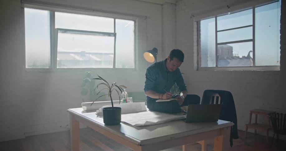 A man is sitting on an office desk writing in a book and flipping through papers on the desk