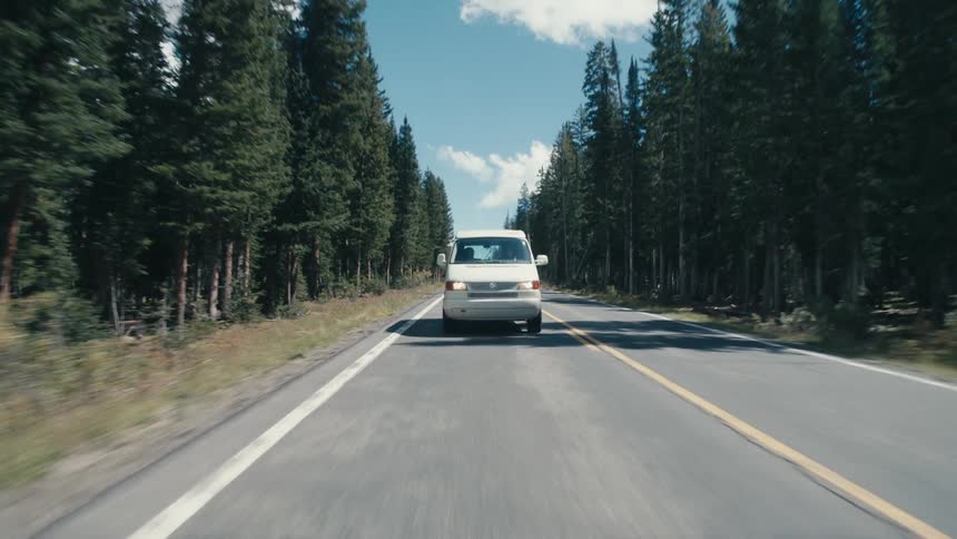 A white van is traveling on a road surrounded by trees