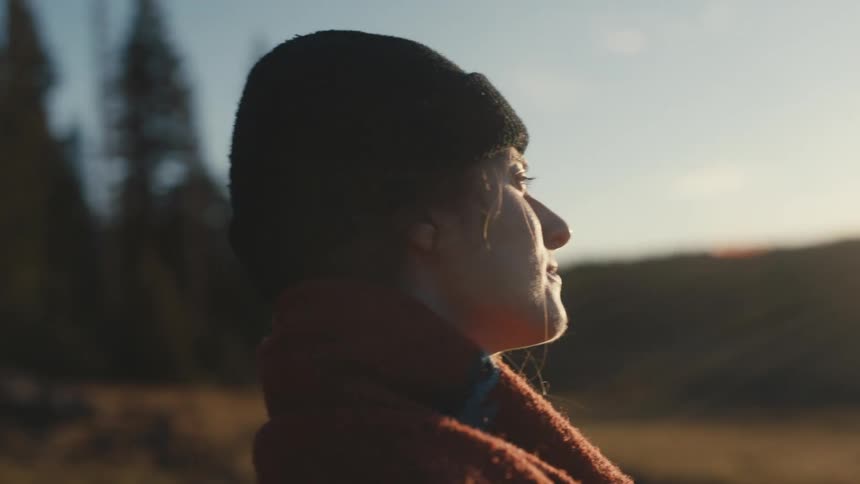 A woman wearing a knit cap is standing outside smiling and the sun is shining brightly behind her