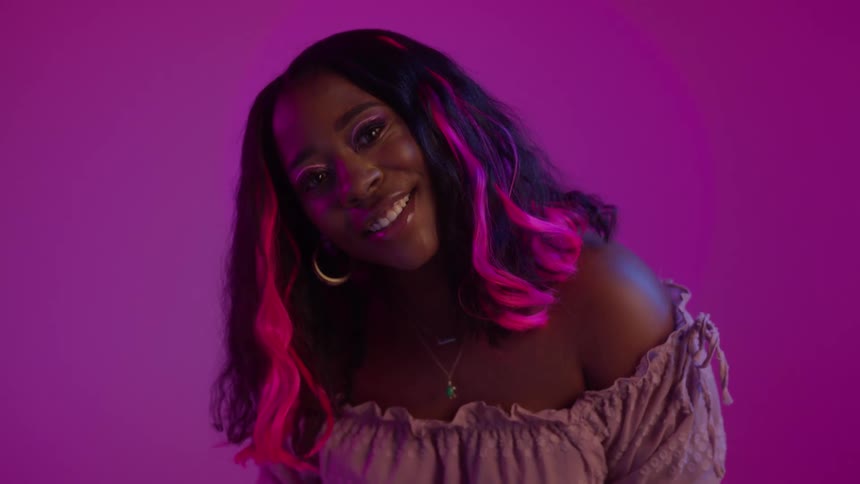 A black woman with pink streaks in her hair is smiling and posing with a hot pink wall behind her
