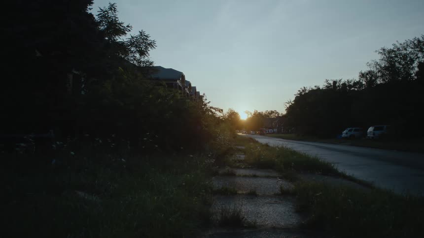 The sun sets at the end of a road with a building and vans and automobiles parked on the roadside near trees