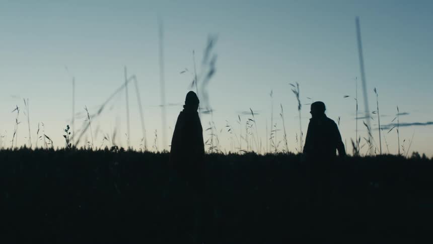 Two women walk out into the grass then stand wrapped in a single blanket to watch the sunrise
