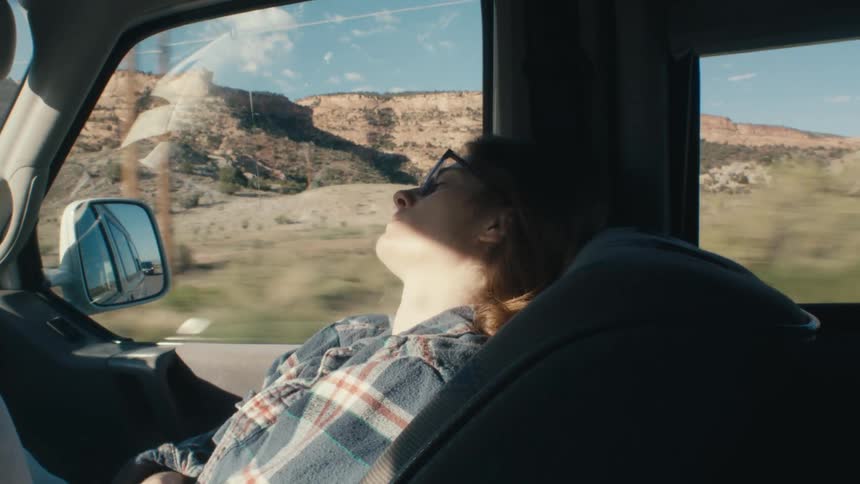 Person sleeps in passengers seat as vehicle travels past scrubland and plateaus