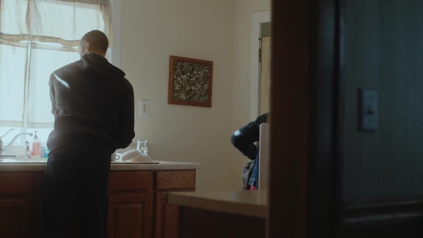 A man is standing at a kitchen sink washing dishes when a woman comes into the kitchen and hands him a paper