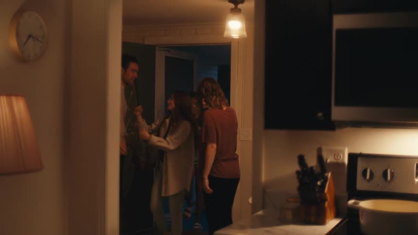 A pregnant woman cooking in a home kitchen stops to answer the door and greet the two young women waiting outside with a hug