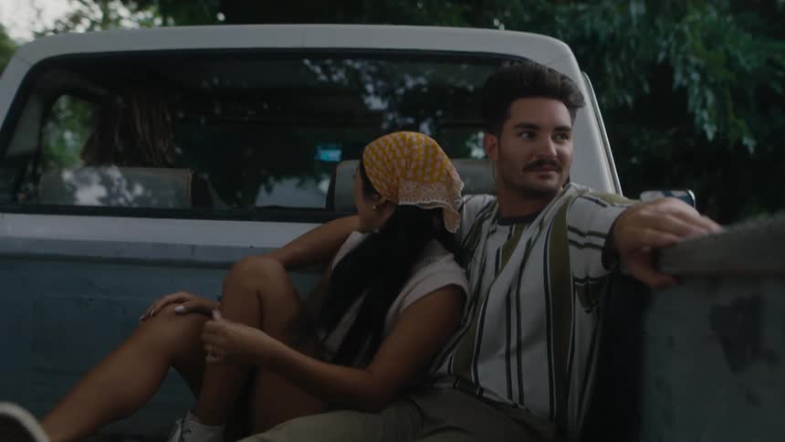 A man and a woman are sitting in the back of a pickup truck as it travels on a road surrounded by trees