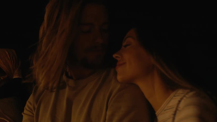 Woman reclines her head on the shoulder of a handsome man sitting near a campfire at night