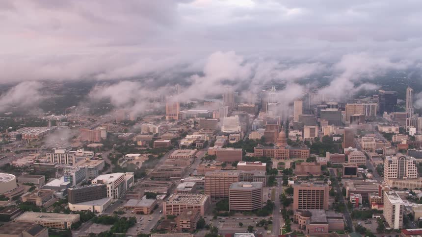 Clouds float low over a city with many buildings and trees as automobiles travel on the roads