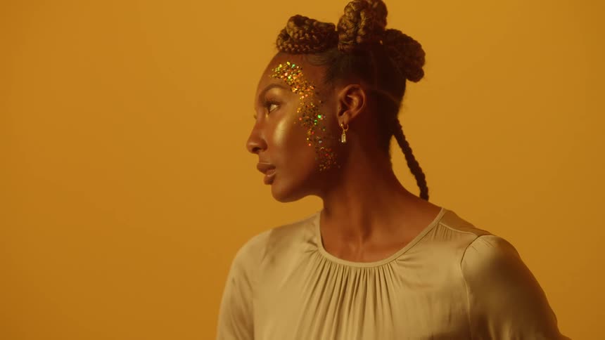 A black woman with glitter on her face is standing near a bright yellow wall