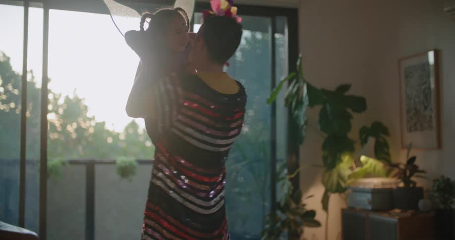 A man is wearing a sequined shirt and a party hat and lifting his young daughter wearing fairy wings in the air