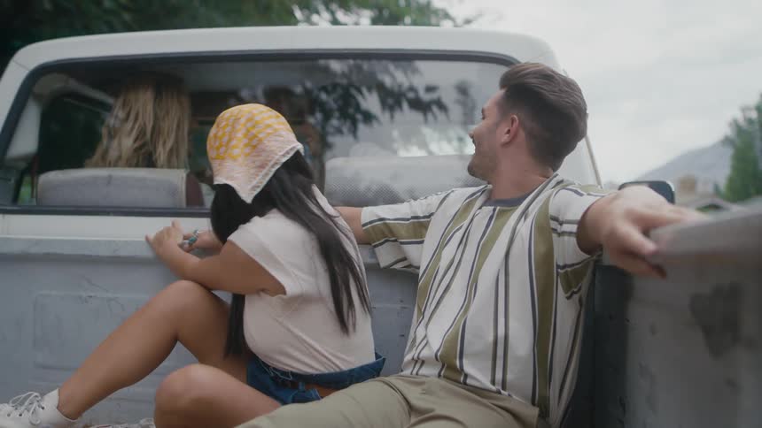A man and a woman are sitting in the back of a pickup truck and she kisses him as the girls in the truck look at them and smile